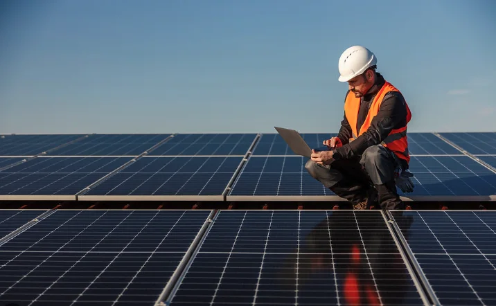 Solar technician in a hard hat and safety vest reviewing a laptop on a solar array